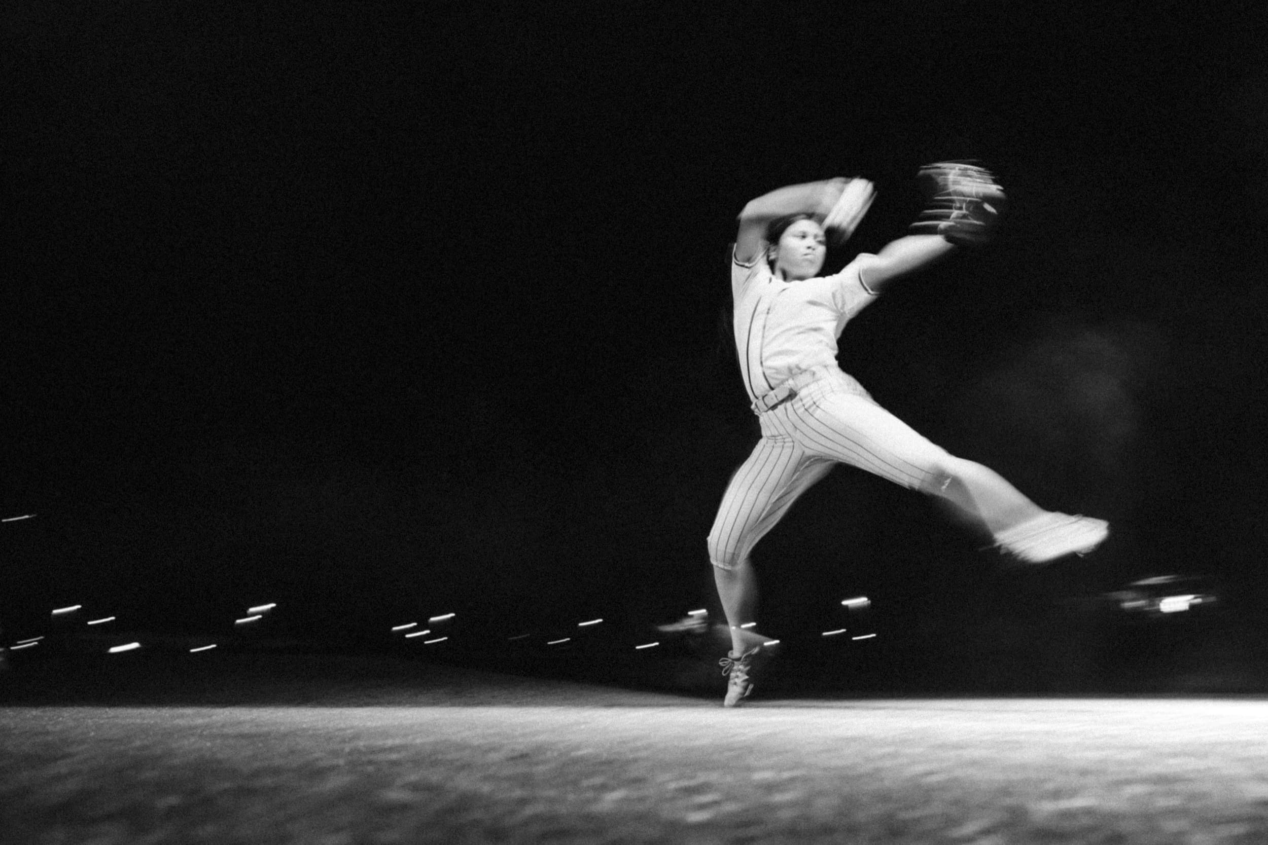 A pitcher mid-delivery under stadium lights, captured with motion blur.